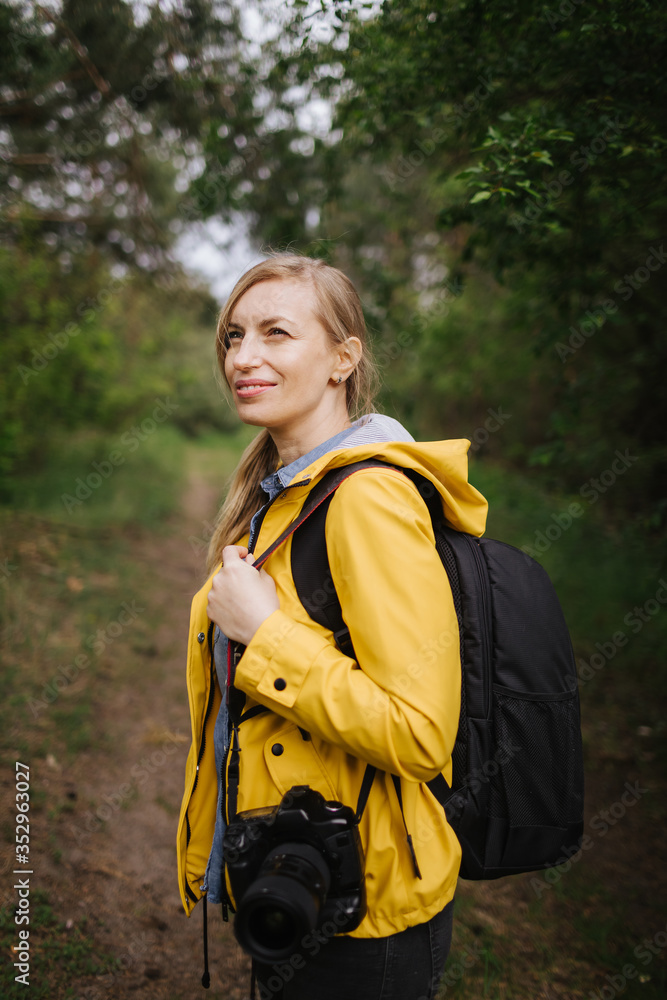 Charming mature woman with blond hair standing among spring forest with big backpack and professional camera. Photographer in yellow jacket working on fresh air Charming mature woman with blond hair standing among spring forest with big backpack and professional camera. Photographer in yellow jacket working on fresh air