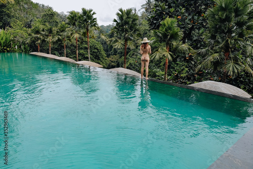Infinity pool at luxurious exotic island. Back view of woman walking on edge of pool and enjoy jungle view wearing beige bikini and hat. Vacation concept