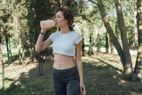 The girl with blond blonde curly hair in a white top and gray leggings rests in the shade after training, drinks water and looks to the side
