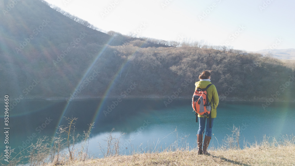 A girl traveler in a coat and a backpack is standing on the shore of the lake. A trip in the off-season. Cold weather