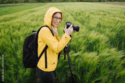 Cheerful nature photographer in yellow jacket and eyeglasses using professional equipment while taking photos of wheat field. Concept of working process and shooting