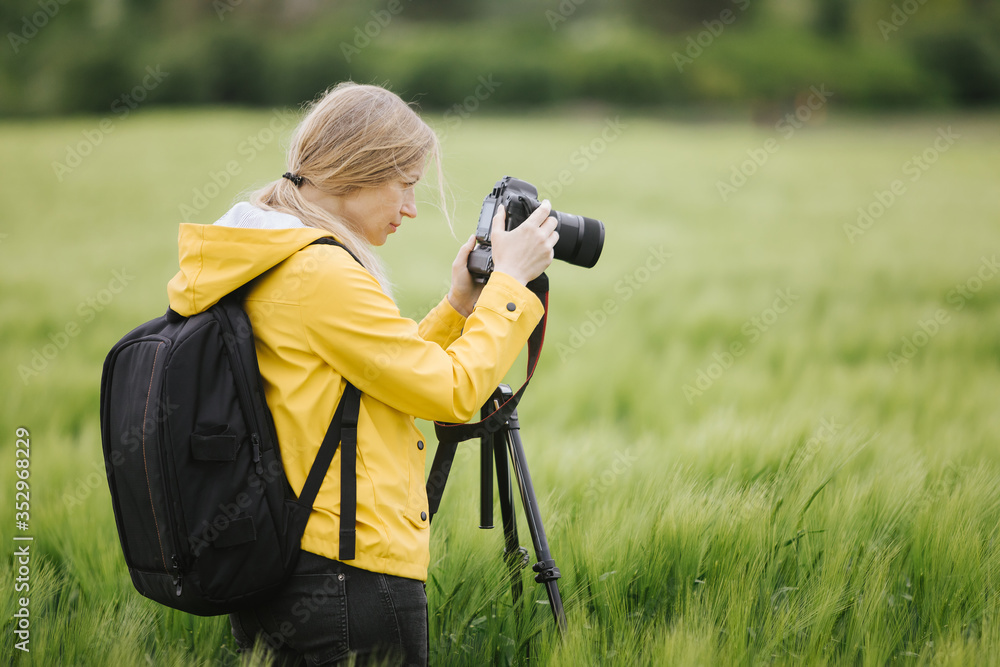 Competent blonde photographer taking pictures of beautiful spring nature. Beautiful woman in yellow jacket standing at green field with tripod and digital camera.