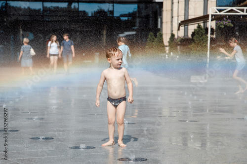 little boy with blond hair walking around the city and enjoying the sunshine. Fountain with water making a rainbow.