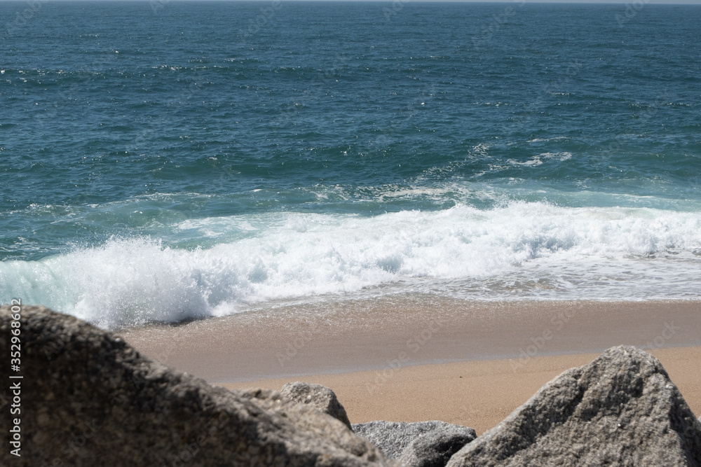 White waves breaking on sandy beach in summer in Portugal with rocks in foreground White waves breaking on sandy beach in summer in Portugal with rocks in foreground