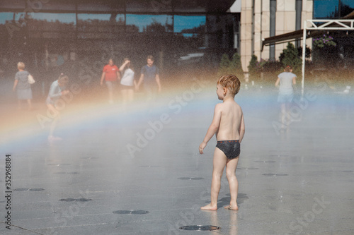 little boy with blond hair walking around the city and enjoying the sunshine. Fountain with water making a rainbow.