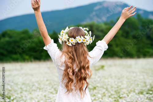 Beautiful young girl with curly hair in chamomile field.  Beautiful girl with chamomile wreath on flowering field in summer. Beautiful curly hair. Freedom concept.