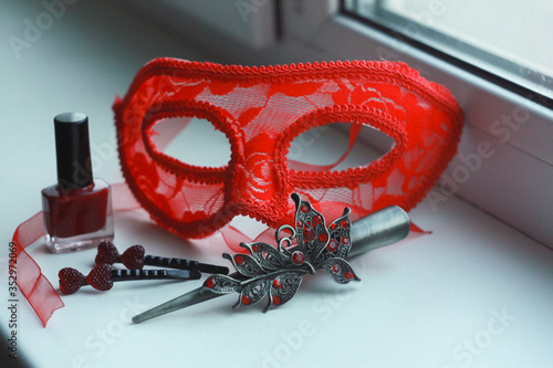 Composition of red vintage carnival mask, nail polish, metal hair clips with rhinestones, pomegranate beads made of natural stone on a white background on the windowsill. photo