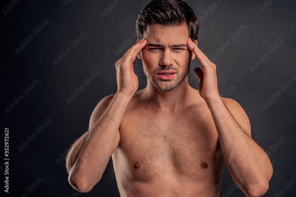 Handsome young bearded man isolated. Portrait of topless muscular man is standing on gray background. Man holding his temple. Experiencing headache Handsome young bearded man isolated. Portrait of topless muscular man is standing on gray background. Man holding his temple. Experiencing headache