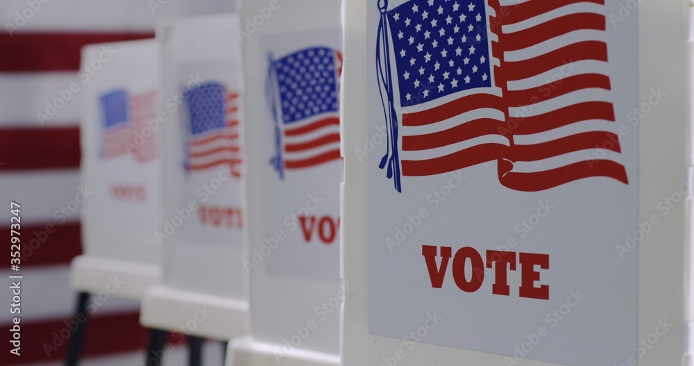 © vesperstock - CU straight on row of voting booths at polling station during American election. US flag in background.