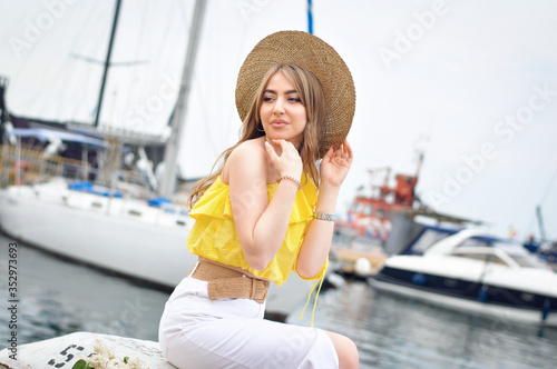 A beautiful young woman with blond curls in a straw hat sits against a white yacht and looks back. 