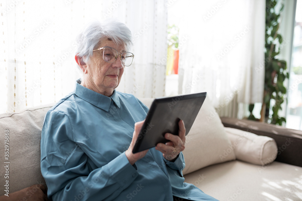 Serious senior gray-haired Caucasian woman sitting on a sofa in brightly lit living room and browsing internet or looking at family photos on a tablet