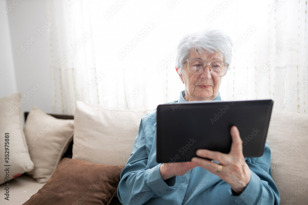 Sad and lonely senior gray-haired Caucasian woman sitting on a sofa in brightly lit living room and browsing internet or looking at family photos on a tablet Sad and lonely senior gray-haired Caucasian woman sitting on a sofa in brightly lit living room and browsing internet or looking at family photos on a tablet