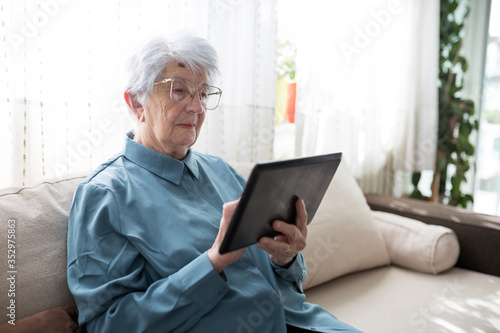 Obraz na płótnie Serious senior gray-haired Caucasian woman sitting on a sofa in brightly lit living room and browsing internet or looking at family photos on a tablet w salonie