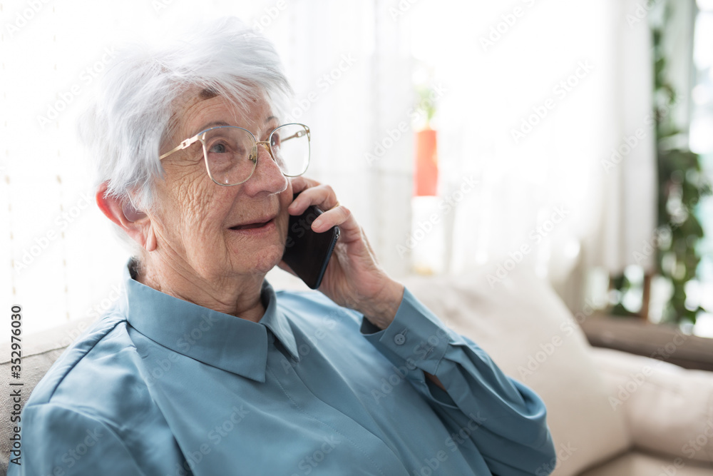 Smiling senior gray-haired Caucasian woman sitting on a sofa in brightly lit living room and talking on the phone Smiling senior gray-haired Caucasian woman sitting on a sofa in brightly lit living room and talking on the phone