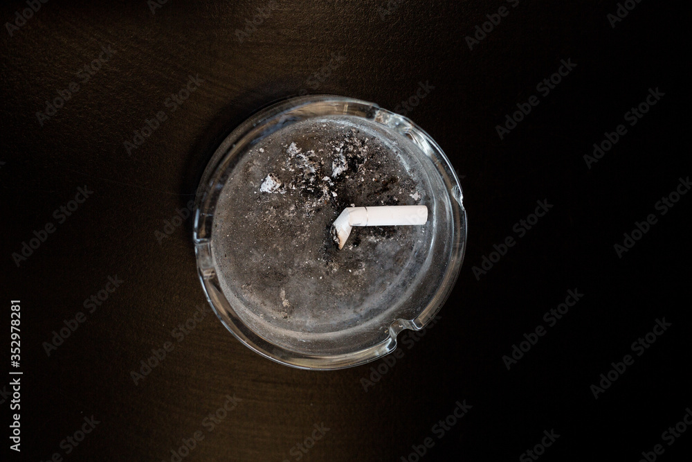 Glass transparent ashtray with one white cigarette butt and ash. Smoking concept. Black background with copy space. Isolation and loneliness in objects. Glass transparent ashtray with one white cigarette butt and ash. Smoking concept. Black background with copy space. Isolation and loneliness in objects.