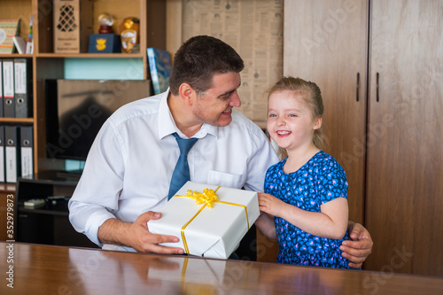 Little beautiful girl blonde gives a gift to her dad in his office at work on father's day. Holiday concept and family values.