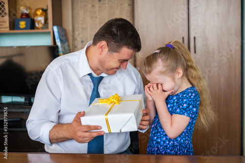 Little beautiful blonde girl is embarrassed when giving a gift to her dad in his office at work on father's day. Holiday concept and family values.