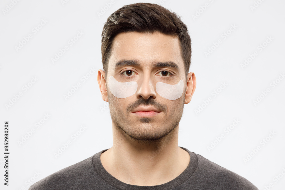 Young male using beige under eye patches to remove traits of exhaustion or sleep deprivation and look fresher, isolated on gray background Young male using beige under eye patches to remove traits of exhaustion or sleep deprivation and look fresher, isolated on gray background