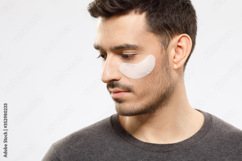 Obraz premium Young male looking aside and down with eye patch preventing from dark circles and wrinkles, taking care of his face skin, isolated on gray background