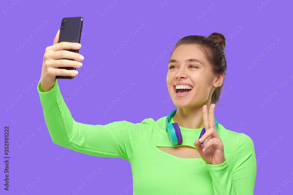 Young happy girl wearing green neon tep and headphones around neck, taking selfie photo with her phone, smiling and showing V sign, isolated on purple backgroound Young happy girl wearing green neon tep and headphones around neck, taking selfie photo with her phone, smiling and showing V sign, isolated on purple backgroound