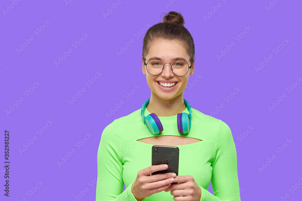 Portrait of young smiling girl, wearing green neon top, round glasses and wireless headphones around neck, holding smartphone in hands, isolated on purple background Portrait of young smiling girl, wearing green neon top, round glasses and wireless headphones around neck, holding smartphone in hands, isolated on purple background