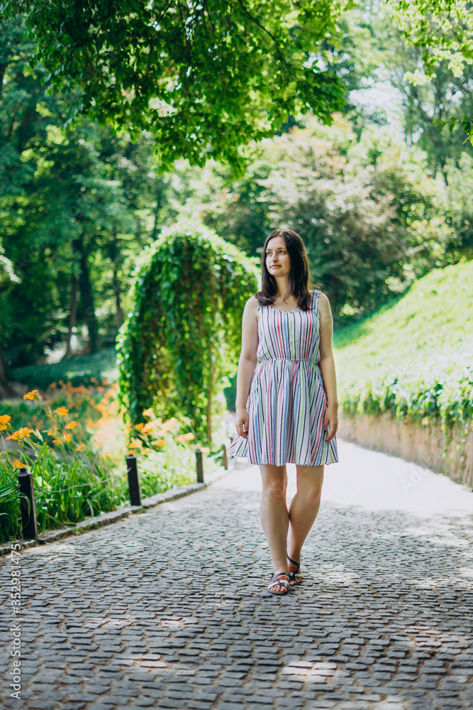 Sofia Park, Uman. The girl walks along the alleys of the park on a sunny day. Brunette girl in a white dress walks in a beautiful landscape park. Path with cobblestones in a beautiful green park. Sofia Park, Uman. The girl walks along the alleys of the park on a sunny day. Brunette girl in a white dress walks in a beautiful landscape park. Path with cobblestones in a beautiful green park.