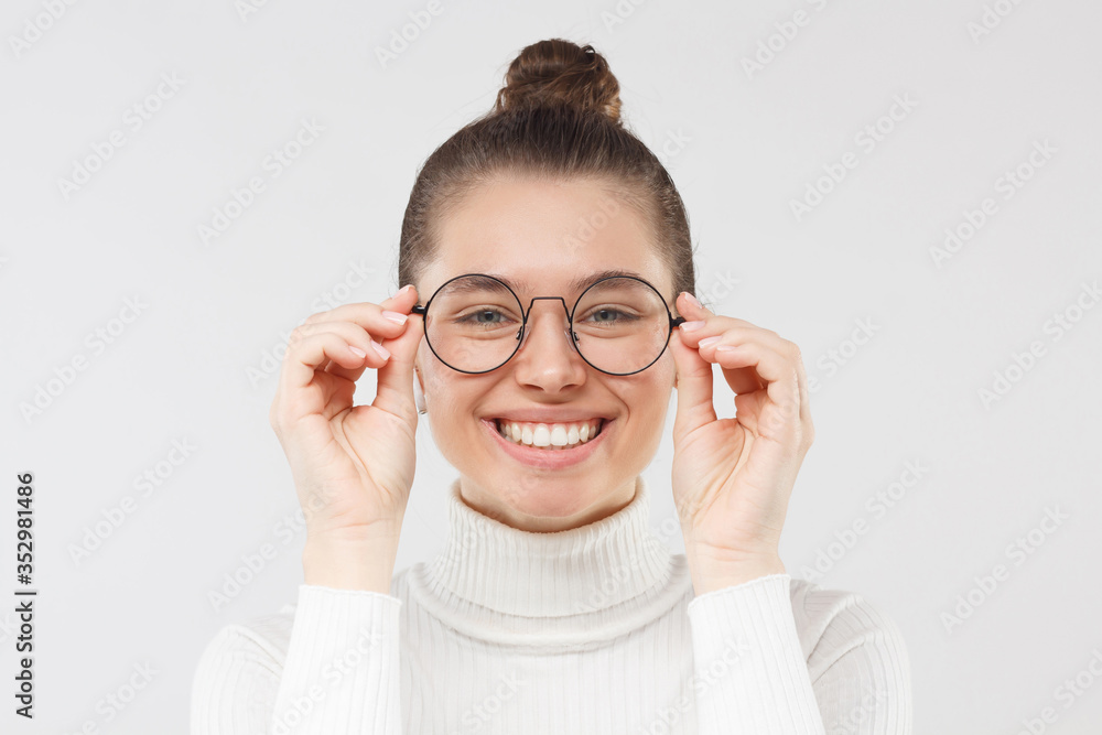 Young smiling woman in white sweater looking at something through round eyeglases with interest and attention, isolated on gray background Young smiling woman in white sweater looking at something through round eyeglases with interest and attention, isolated on gray background
