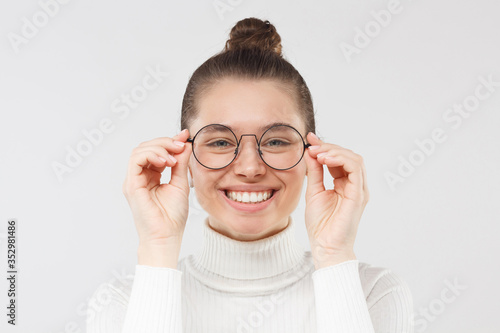 Young smiling woman in white sweater looking at something through round eyeglases with interest and attention, isolated on gray background photo