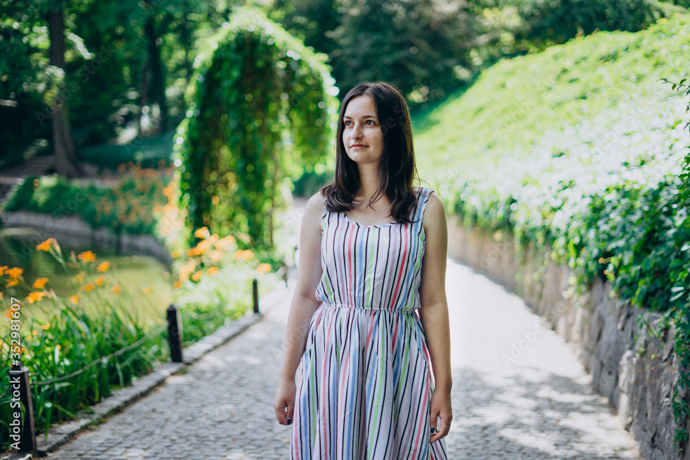 Sofia Park, Uman. The girl walks along the alleys of the park on a sunny day. Brunette girl in a white dress walks in a beautiful landscape park. Path with cobblestones in a beautiful green park. Sofia Park, Uman. The girl walks along the alleys of the park on a sunny day. Brunette girl in a white dress walks in a beautiful landscape park. Path with cobblestones in a beautiful green park.