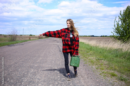 An attractive young blonde girl, in a red shirt, stands in the middle of the road with a canister, outstretched a hand to stop the car and borrow a few liters of fuel. Windy spring day.