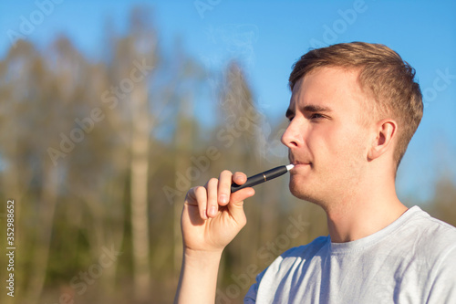 young man smokes, puffs an electronic cigarette against background of nature. Handsome guy with tobacco heating system, vaping in the fresh air. bad habit. copy space, place for text
