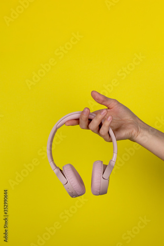 A young woman holds a pair of pink wireless headphones in her hand. Hand of a girl with headphones on a yellow background. Musical concept.