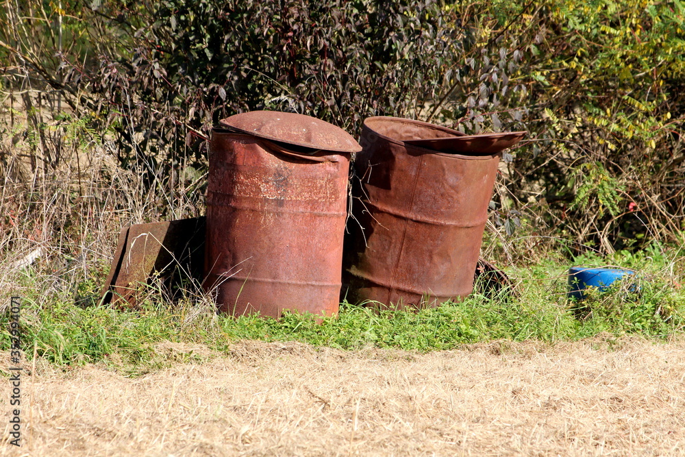 Two completely rusted beaten old oil barrels left as illegal garbage in nature next to other metal junk surrounded with uncut grass and dense trees in background on warm sunny autumn day
