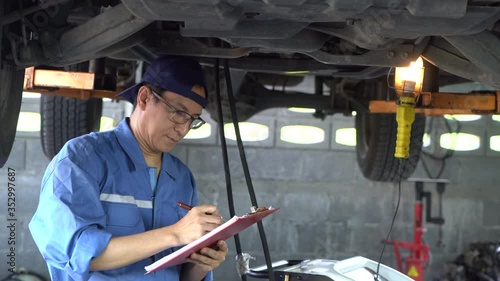 Asian Man mechanic inspection writing note on clipboard insurance notepad with car engine.for transport automobile automotive image.