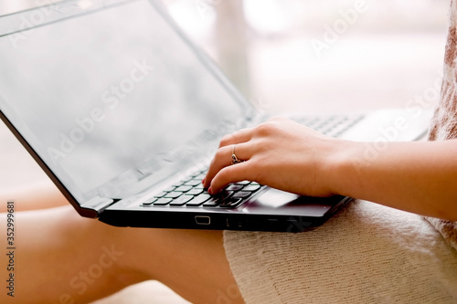 girl in a light knitted sweater sits near the window and holds a laptop. Female hands print on a notebook, remote work at home.