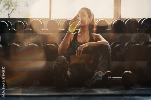 caucasian woman in sportswear sitting on floor near dumbells rack resting and rekaxing after having excercise and workout in gym and fitness club with sun flare, selective focused photo