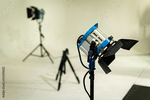 Old vintage fresnel light on tripod in a film, television or photography studio. It's gray and blue. There's another one in the back; both are turned off. White and empty filming set. photo