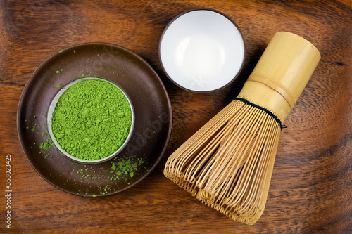 Powdered matcha tea in a cup and green leaves with bamboo matcha tea whisk also know as chasen on a wooden background.