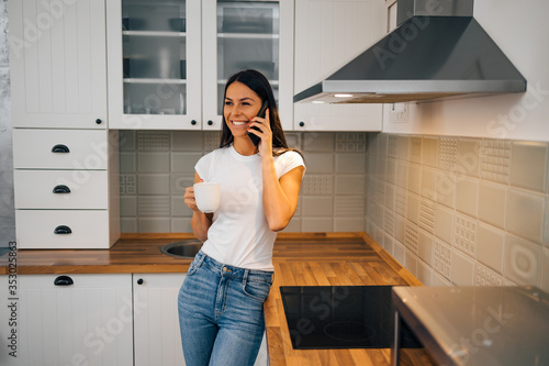 Happy young woman talking on mobile phone while standing with a cup of tea in the kitchen, portrait.