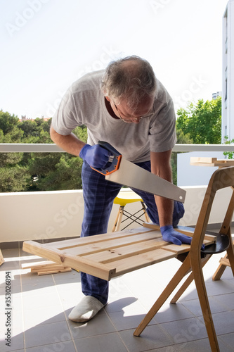 Mature man with saw cutting timber on home balcony, assembling or repairing wooden furniture, doing carpentry work. Vertical full length shot. Lockdown activity concept