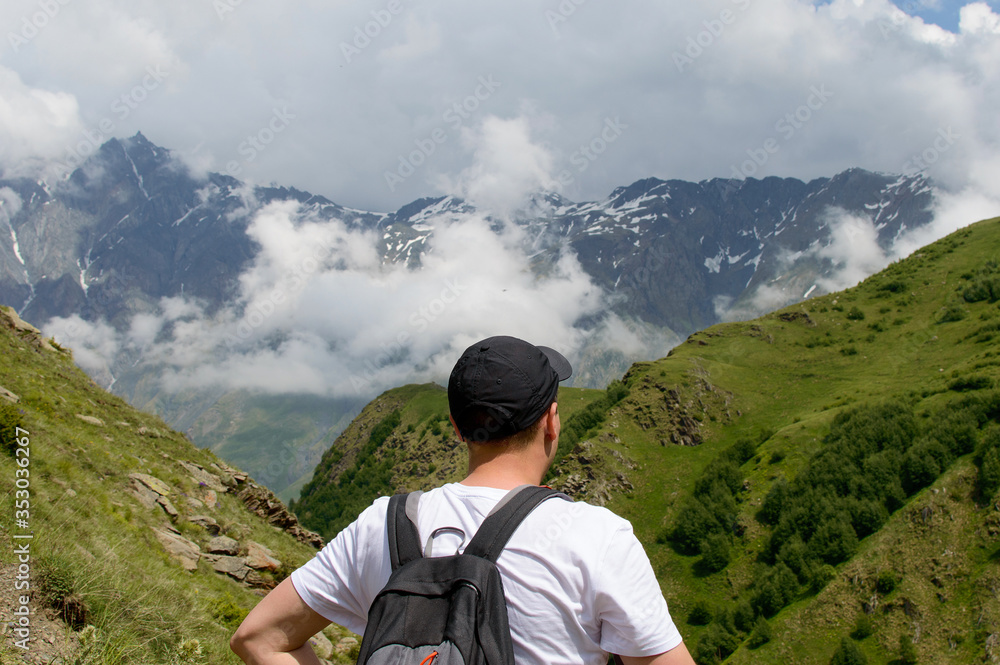 A young man looks in mountains in a canyon. Back of the man in white t-shirt with backpack a cloudy green hills. Tourist hikes on a top of the mountain