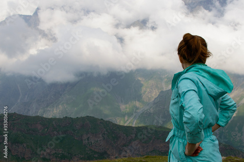 Beauty redhead girl looking a sunrise on mountains in a canyon. Back of the woman looking a cloudy green hills. Handsome lady in light cloak in the top of a mountain. Tourist enjoy a moment in nature.