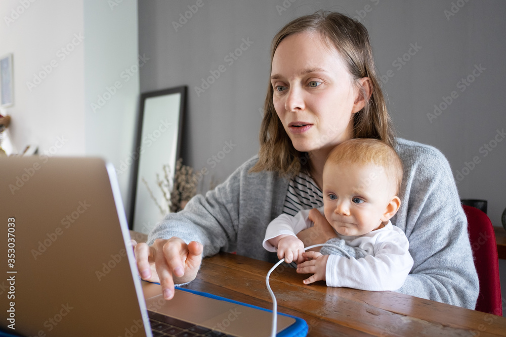 Cute baby holding laptop cable and watching for mother. Woman holding daughter and working on computer at home. Online connection and staying at home on isolation concept Cute baby holding laptop cable and watching for mother. Woman holding daughter and working on computer at home. Online connection and staying at home on isolation concept