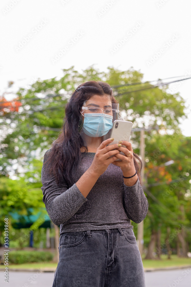 Young Indian woman with mask and face shield using phone at the park outdoors Young Indian woman with mask and face shield using phone at the park outdoors