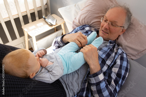 Cute red-haired baby laying on grandpa belly and carefully listening him. Senior man holding grandchild legs and talking with cute toddler. Help from grandparents and staying at home concept