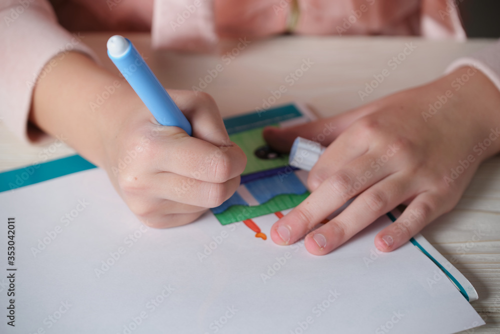 A girl in a pink sweatshirt draws with a light blue felt-tip pen at a table. Blurred front background. Beige table A girl in a pink sweatshirt draws with a light blue felt-tip pen at a table. Blurred front background. Beige table