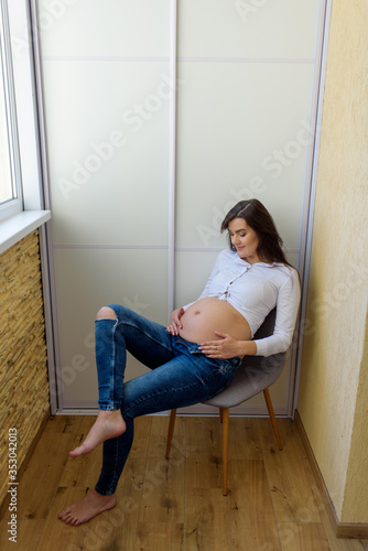 Beautiful pregnant girl is sitting on a chair on the balcony. The girl stretched her stiff legs. Self-isolation.