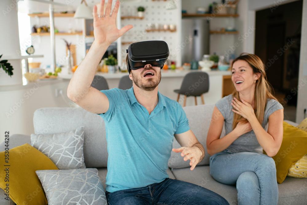 Smiling young man using VR headset at home on couch. Man and his wife enjoying virtual reality at her apartment. Smiling young man using VR headset at home on couch. Man and his wife enjoying virtual reality at her apartment.