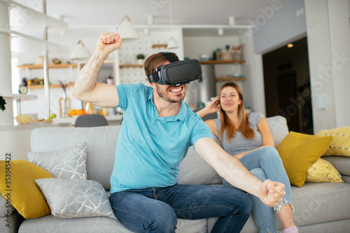 Smiling young man using VR headset at home on couch. Man and his wife enjoying virtual reality at her apartment. 