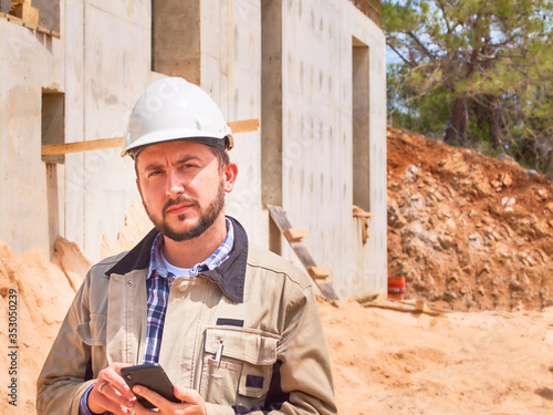 Caucasian bearded civil engineer or construction worker in white hardhat uses his pnone opposite construction site photo